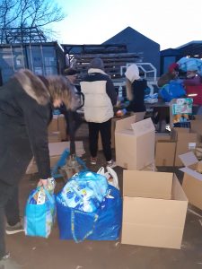 Pakowanie darów dla mieszkańców Ukrainy. Na zdjęciu cztery osoby - jedna z nich trzyma za rączki wypełnioną, plastikową torbę. Na zdjęciu porozstawiane na ziemi kartony i torby z przekazanymi darami.