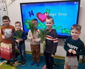 Chłopcy z oddziału przedszkolnego stoją obok siebie na tle tablicy, na której na kolorowym tle wyświetla się napis Miłego Dnia. Chłopcy trzymają w dłoniach czerwone tulipany oraz torby z prezentami.
