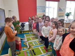 Dziewczynki z oddziału przedszkolnego stoją obok siebie. Na przeciw stoją chłopcy trzymający czerwone tulipany i torbę z prezentem.