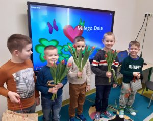 Chłopcy z oddziału przedszkolnego stoją obok siebie na tle tablicy, na której na kolorowym tle wyświetla się napis Miłego Dnia. Chłopcy trzymają w dłoniach czerwone tulipany oraz torby z prezentami.