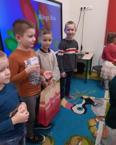 Czterech chłopców z oddziału przedszkolnego stoi obok siebie w rzędzie na tle tablicy interaktywnej, na której na kolorowym tle wyświetla się napis Miłego Dnia. Jeden z chłopców trzyma w dłoni papierową torbę na prezent.