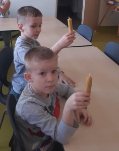 Dwóch chłopców siedzi przy stoliku. W dłoniach trzymają świece wykonane z wosku pszczelego.