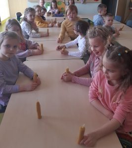 Dzieci siedzą przy połączonych stolikach, na których stoją świeczki wykonane z wosku pszczelego. Większość świeczek dzieci trzymają w dłoniach.
