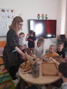 Pani stoi przy stoliku, na którym zgromadzono przedmioty związane z pszczelarstwem. W okół stolika stoją dzieci słuchające Pani.