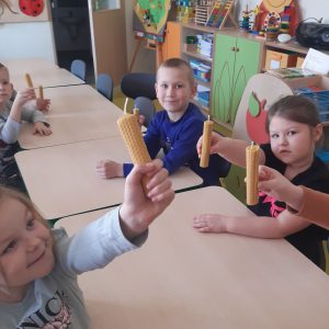 Dzieci siedzą przy połączonych stolikach. W wyciągniętych dłoniach trzymają świeczki wykonane z wosku pszczelego.