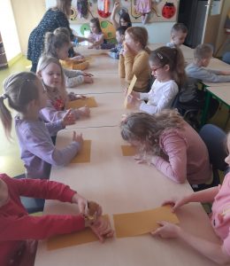 Dzieci siedzą przy stoliku. Trzymają w słoniach prostokątne kawałki wosku pszczelego, z których zrobią świeczki.