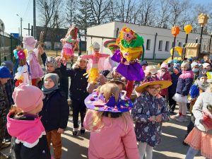 Uczniowie oddziału przedszkolnego stoją na placu przed szkołą. W rękach trzymają marzanny a na głowachmają kolorowe kapelusze specjalnie wykonane na pierwszy dzień wiosny.