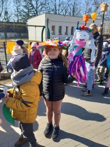 Dzieci stoją na dworze. Na pierwszym planie dziewczynka trzyma w lewej ręce marzannę.
