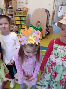 Uczennice pozują do zdjęcia w kapeluszach ozdobionych kwiatami. Dziewczynka stojąca na pierwszym planie pochyla głowę tak by jej kapelusz był lepiej widoczny.