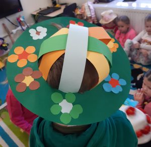 Zdjęcie głowy ucznia w zielonym kapeluszu. Kapelusz wykonany jest z papieru i ozdobiony kolorowymi kwiatami. Po środku umieszczone są kolorowe, szerokie paski papieru, pomiędzy którymi widoczne są włosy ucznia.