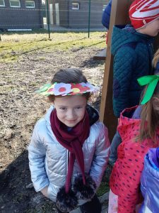 Uczennica pozuje do zdjęcia w papierowym nakryciu głowy. Kapelusz ma szerokie rondo ozdobione papierowymi kwiatami oraz wolną przestrzeń na czubku przez którą wystaje głowa dziewczynki.