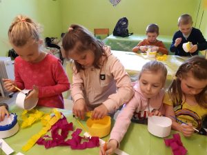 Uczniowie oddziału przedszkolnego ozdabiają białe miseczki kolorowym papierem. Dzieci pracują przy stołach pokrytych zielonym materiałem i ustawionych w dwóch rzędach.