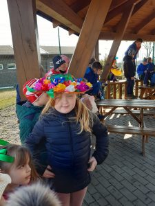 Uczennica pozuje do zdjęcia w kolorowym kapeluszu przybranym papierowymi kwiatami. W tle widać drewnianąkonstrukcję altany pod którą stoi uczennica oraz inne dzieci.