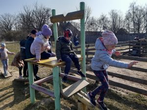 Uczniowie oddziału przedszkolnego wspinają się na duże drewniane krzesło ustawione obok zagrody na podwórku babaluby. Dwoje uczniów siedzi na krześle a ich koleżanka z niego schodzi po drewnianych palikach zamontowanych pod skosem z jednej strony krzesła.