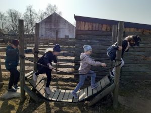 Uczniowie oddziału przedszkolnego przechodzą przed drewnianą kładkę opadającą aż do ziemi. W tle widać drewniane zabudowy.