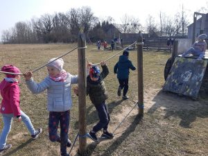 Uczniowie oddziału przedszkolnego bawią się na podwórku przed ośrodkiem babaluba. Dzieci na pierwszym planie chodzą po linie przypomocowanej do drewnianych palików tuż nad ziemią. Dzieci trzymają się rękoma drugiej linki zamocowanej na tych palikach na wysokości ich głów.