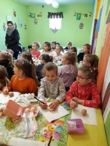 Dzieci z oddziału przedszkolnego siedzą przy stołach zakrytych zielonymi obrusami. Na stołach leżą wytłaczanki z jajkami. Przed uczniami na białych serwetkach leżą jajka. W tle widać przechodzącego nauczyciela oraz zielone ściany pomieszczenia.