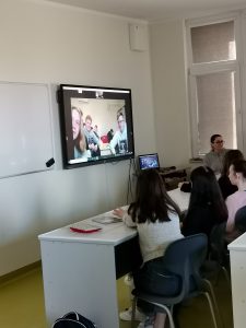 Uczniowie siedzą w rzędzie przy białym stole. Przed nimi na ścianie wisi zamontowany ekran, na którym widać innych uczniów.