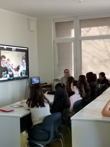 Uczniowie siedzą w rzędzie przy białym stole. Przed nimi na ścianie wisi zamontowany ekran, na którym widać innych uczniów.