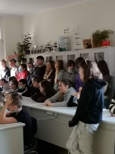 Uczniowie siedzą w dwóch rzędach białych ławek. Za ich plecami opierając się plecami o regał stoją w rzędzie ich koledzy. Wszyscy patrzą w jedną stronę.