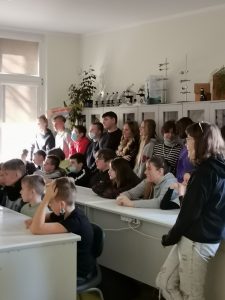 Uczniowie siedzą w dwóch rzędach białych ławek. Za ich plecami opierając się plecami o regał stoją w rzędzie ich koledzy. Wszyscy patrzą w jedną stronę.