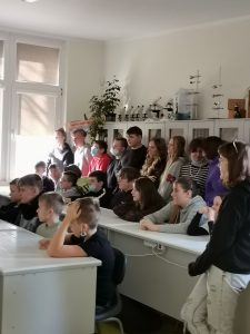 Uczniowie siedzą w dwóch rzędach białych ławek. Za ich plecami opierając się plecami o regał stoją w rzędzie ich koledzy. Wszyscy patrzą w jedną stronę.