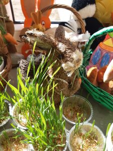 Zdjęcie dekoracji wielkonocnej. Za małymi doniczkami z zieloną trawą umieszczono zajączka wykonanego z kolorowanego siana. W tle widać koszyczki z pisankami.