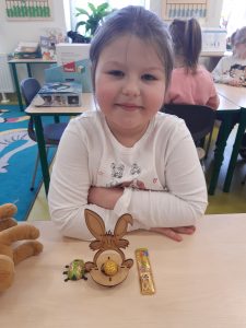 Uczennica siedzi przy stoliku o który opiera swoje przedramiona. Przed nią ustawiony jest model zajączka oraz czekolaadowy żuk i zajączek w pazlotkach.