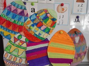 Na białej tablicy przyczepione wycięte z papieru pisanki ozdobione na kolorowo przez uczniów.