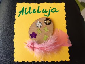 Zdjęcie świątecznej kartki - na żółtym papierze naklejone brązowe jajko ozdobione wklejonymi motylkami i gwiazdkami. Pod jajkiem przymocowano różowe piórko a nad dekoracją zielony napis Alleluja.
