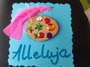 Zdjęcie świątecznej kartki - na niebieskim papierze naklejone brązowe jajko ozdobione wklejonymi motylkami i diamentami. Obok jajka różowe piórko a pod spodem niebieski napis Alleluja.