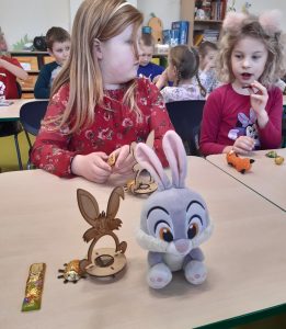 Dwie uczennice siedzą obok siebie przy złączonych na stołach. Jedna z nich je czekoladkę, druga ją obserwuje. Na stoliku przed dziewczynkami leży pluszowa maskotka oraz złożony model.