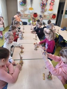 Uczniowie oddziału przedszkolnego siedzą przy połączonych stolikach. Przed dziećmi stoją modele króliczków. Uczniowie przyglądają się swoim modelom.