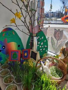 Wielka dokoracja okna oddziału przedszkolnego, na parapecie którego ustawiono małe doniczki z zieloną trawą koszyczek z pluszowym zajączkiem, kartki świąteczne z narysowanymi pisamkami oraz gałązki z doczepionymi papierowymi żonkilami wystające z srebrnego termosu.
