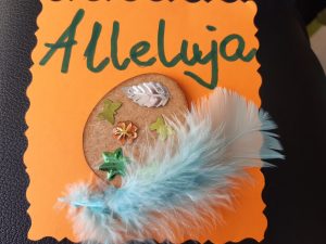 Zdjęcie świątecznej kartki - na pomarańczowym papierze naklejone brązowe jajko ozdobione wklejonymi motylkami, listkiem i gwiazdką. Obok jajka jasnoniebieskie piórko a nad dekoracją zielony napis Alleluja.