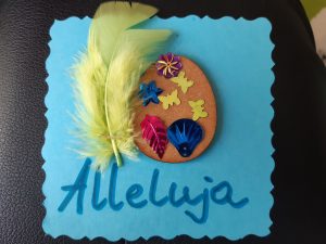 Zdjęcie świątecznej kartki - na niebieskim papierze naklejone brązowe jajko ozdobione wklejonymi motylkami, listkiem i muszelką. Obok jajka zielone piórko a pod spodem niebieski napis Alleluja.