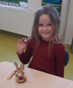 Uczennica siedzi przy stoliku. Trzyma w ręku czekoladkę a przed nią stoi model zajączka. Dziewczynka się uśmiecha.