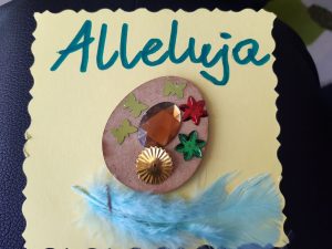 Zdjęcie świątecznej kartki - na żółtym papierze naklejone brązowe jajko ozdobione wklejonymi motylkami, gwiazdkami i diamentem. Pod jajkiem przymocowano niebieskie piórko a nad dekoracją zielony napis Alleluja.