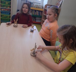 Trzy dziewczynki siedzą przy stoliku. Przed nimi na stole ustawiono modele zajączków. Jedna z dziewczynek składa swój model.
