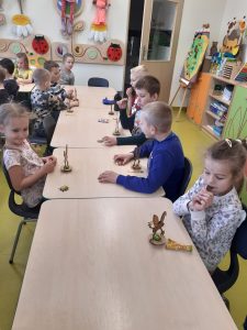 Uczniowie oddziału przedszkolnegosiedzą przy połączonych stołach. Przed dziećmi stoją modele zajączków a dzieci jedzą czekoladki.