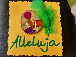 Zdjęcie świątecznej kartki - na żółtym papierze naklejone brązowe jajko ozdobione wklejonymi motylkami, listkiem i diamentami.Obok jajka zielione piórko a pod spodem zielony napis Alleluja.