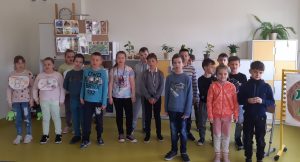 Uczniowie stoją obok siebie na tle kolorowej meblościanki. Dwie dziewczynki po lewej stronie mają zawieszone na szyi owalne kartki z kolorowymi literkami B i E.