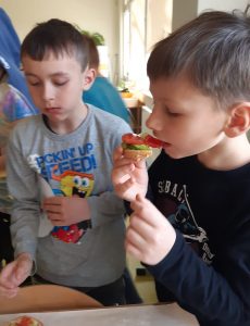 Dwóch uczniów stoi obok siebie. Jeden z nich je zdrową kanapkę z pomidorem a drugi właśnie kończy jeść swoją przekąskę.