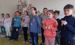 Uczniowie stoją obok siebie pod ścianą sali. Są w trakcie recytowania wiersza. Część z nich ma zamknięte oczy.