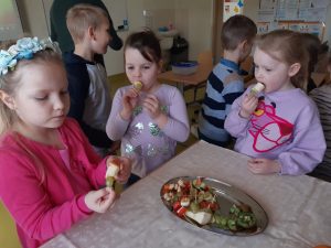 Trzy dziewczynki stoją obok siebie przy narożniku stołu. Dziewczynki jedzą koreczki. Na stole przyktytym białym obrusem stoi półmisek z pozostałymi koreczkami. W tle widać chodzących po klasie uczniów.