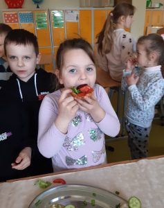 Uczennica na pierwszym planie je kanapkę z pomidorem i ogórkiem. Dziewczynka stoi przy stole na którym leży półmisek. W tle widać inne dzieci chodzącepo klasie.
