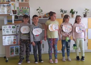 Pięcioro uczniów stoi na tle biało-żółtej meblościanki z półkami i szafkami. Dzieci mają zawieszone na szyi owalne kartki każda z inną literą: F, D, C, B, E.