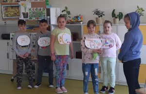 Sześcioro uczniów stoi w klasie na tle meblościanki z półkami i szafkami. Piątka dzieci ma zawieszone na szyi kartki w owalnym kształcie z kolorowymi literkami - F,D,C,B, i E. Każde dziecko ma zawieszoną kartkę z inna literką. Uczennica bez literki stoi naprzeciwko kolegów.