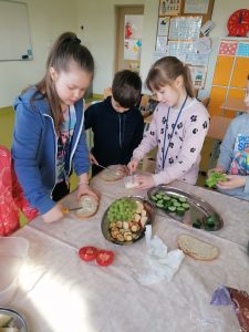 Uczniowie w trakcie przygotowywania zdrowego posiłu. Troje dzieci stoi przy stole zakytym białym obrusem. i smaruje kawałki chleba masłe. Na stole stoją dwie tace z kolorowymi dodatkami do kanapek.