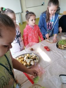 Uczniowie przygotowują zdrowe kanapki. Dzieci stoją przy stole zakrytym białym obrusem, na którym ustawiono dwa półmiski z dodatkami do kanapek. Dzieci smarują kawałki chleba masłem i układają na nich sałatę.
